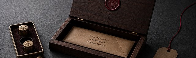 [Translate to English:] Ein hölzerner Briefkasten-Deckel auf glattem Beton mit feinem Riss im präzisen Wachssiegel; im Inneren ein unbeschädigter Briefumschlag, daneben ein Messing-Stempel-Set mit einem leicht verrutschten Stempel und eine Kraftpapier-Etikette mit oxblutfarbenem Faden im kühlen Nordlicht.