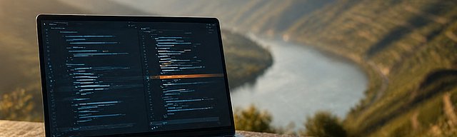 [Translate to English:] Laptop auf Stein im Weinberg, Code-Diff mit warmer Markierung, Morgenlicht.