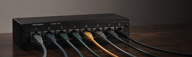 A modern matte-black ethernet patch panel on a dark wooden desk, with several short patch cables in muted colours fanning out in different directions, lit by soft studio light.