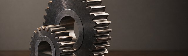 Two interlocking precision-machined steel gears of different sizes standing upright on a dark wooden desk, in soft studio light with a warm rim highlight on the teeth.