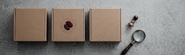 [Translate to English:] Drei identische Kraftpapier-Versandkartons in einer Reihe auf Beton, der mittlere mit gebrochenem dunkelrotem Wachssiegel, daneben ein Messingstempel und eine Lupe.