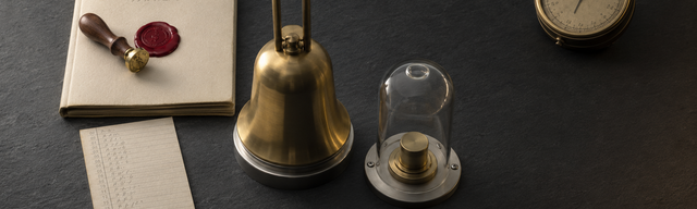 A still life on a matt, dark slate surface, serving as a duty desk in the cyberbunker: at the centre, a burnished brass-coloured signal bell stands upright on a small stainless-steel base, its clapper pointing upwards. To the left of the bell lies a cream-coloured, wax-sealed run book on the slate surface; on the cover is a small embossed round mark and a freshly applied wax seal in a deep, rich oxblood lacquer — the only saturated colour in the picture — with a walnut-handled brass seal head still resting slightly askew on the wax. To the right of the bell, a cylindrical glass bell covers a simple burnished brass push-button actuator, mounted on a stainless steel base plate: the alarm under glass, untouched. At the bottom left lies a narrow cream-coloured duty roster with a column of illegible pencil abbreviations in hourly intervals, held in place by a brass side clip. At the top right of the negative space, a burnished marine chronometer rests at a slight angle on the slate surface, hands on standby. Cool studio key light from the top left, soft warm rim light from the bottom right; the background fades into slate grey and near-black at the right edge of the frame, leaving space for the title overlay. 