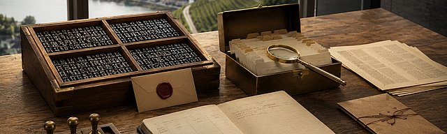 [Translate to English:] Walnuss-Schreibtisch mit antiquem Setzkasten (vier Fächer voller Bleilettern), geöffneter Brass-Karteikassette mit Kraftpaper-Tags, messingfarbener Lupe und einem aufrecht gestellten Oxblood-Wachssiegel-Etikett; im Hintergrund die Glasfront eines modernen Moselhauses mit sonnigem Weinberg-Hang — visuelle Metapher für die vier Schichten eines AI-ready CMS: strukturierte Inhalte, semantische Auslieferung, Agent-Interaktion und Vertrauen.