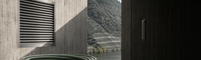 [Translate to English:] Halb geöffnete Betontür eines modernen Rechenzentrums — davor ein dunkles Slate-Plinth mit einem aufgerollten oxblood-Kabel und einem Rack-Modul mit Kraft-Paper-Etikett, im Hintergrund die Mosel-Weinberghänge.