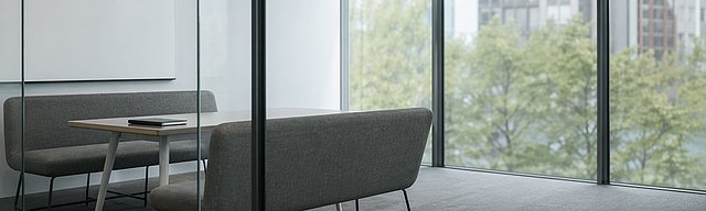 [Translate to English:] Aufgeräumter moderner Besprechungsraum hinter einer Glaswand mit leerer Whiteboard-Wand und zwei gegenüberstehenden Sitzbänken.
