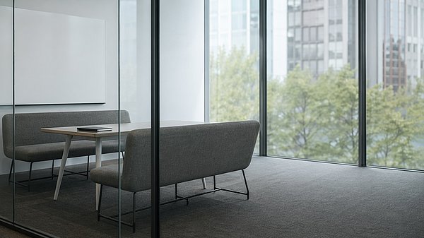 [Translate to English:] Aufgeräumter moderner Besprechungsraum hinter einer Glaswand mit leerer Whiteboard-Wand und zwei gegenüberstehenden Sitzbänken.