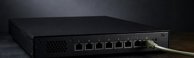 A matte-black server-edge appliance with eight network ports — only one connected with a sage-green patch cable, seven deliberately unused, cool studio light from the left — metaphor for deterministic isolation as security default.