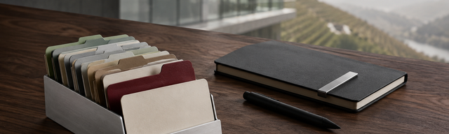 Brushed-aluminium card holder with a fan of pastel-toned index cards on a walnut desk in front of a bright Mosel glass facade; one oxblood-coloured card is pulled forward, alongside a closed matte-black hardcover notebook.