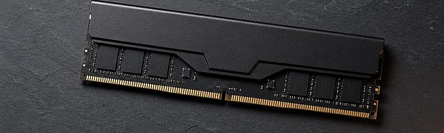 Matte black DDR5 memory module placed diagonally on a dark slate surface, lit by a cool light from the upper left, with visible gold contacts and a short shadow cast to the right.
