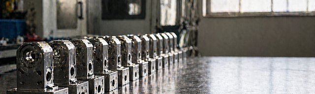 [Translate to English:] Reihe identisch gefertigter Metallbauteile exakt ausgerichtet auf einer Werkbank im Seitenlicht.
