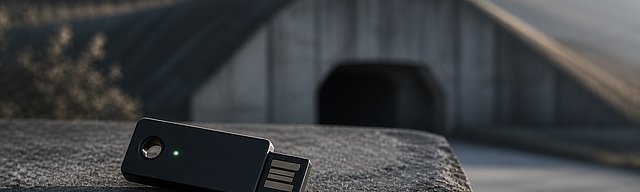 Hardware security token on a weathered concrete plinth, a softly rendered cyber bunker entrance in the Mosel hillside in the background, cool morning light.
