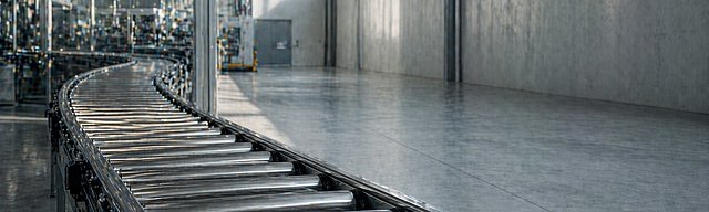 [Translate to English:] Industrielles Förderband mit Rollen in leichter Bewegungsunschärfe in einer hellen Fabrikhalle.