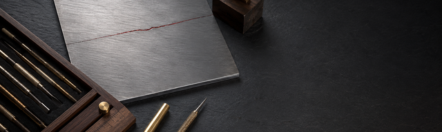 A hardened, brushed-steel inspection plate on matte dark slate; across the plate runs a fine hairline fault, traced with a single thread of oxblood pigment — the discovered vulnerability and the only saturated colour in the image. Lower left, a fitted walnut case of fine probes and engraver's burins in brass and steel; two of them identical — one with a snug brass guard cap, the other bare beside it. Next to it a slim walnut ruler. Upper right, a raised brass loupe on a walnut block, its lens angled diagonally at the hairline fault. Cool studio key light from the upper left; a faint warm rim light from the lower right warms the walnut. The right edge of the frame fades as slate-grey negative space into near-black for a later title overlay.