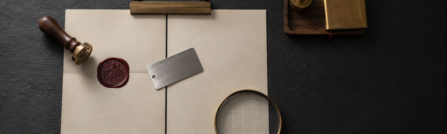 Editorial still life on matte dark slate: two cream paper sheets joined by a brushed brass writing clip. The left sheet bears a still-warm deep oxblood wax impression with an illegible engraved register glyph; a small walnut-and-brass notarial seal press has just been lifted from it. The right sheet appears blank, but a brass cartographer's magnifier on its lower right corner reveals a faint, almost invisible watermark grid against the light. Cool studio key light from the upper left, faint warm rim from the lower right; the right third of the frame is near-black negative space. The only saturated colour is the warm oxblood wax mark on the left sheet.