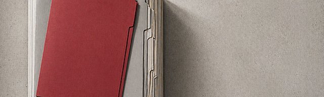 [Translate to English:] Präzise gestapelte graue Aktenmappen von oben fotografiert, eine rote Mappe hebt sich deutlich ab.