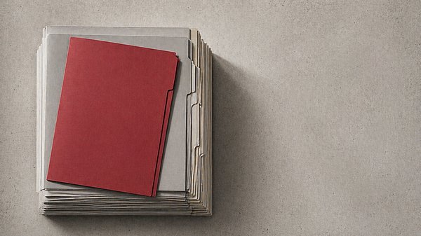 [Translate to English:] Präzise gestapelte graue Aktenmappen von oben fotografiert, eine rote Mappe hebt sich deutlich ab."