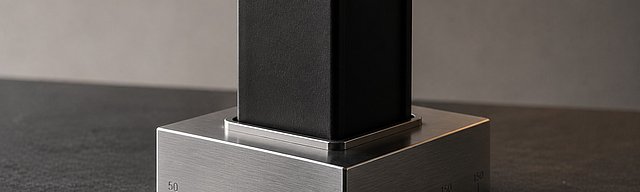 A precision-machined stainless steel foundation block on a dark slate surface, a matte black steel beam rising vertically from its centre, cool directional daylight.